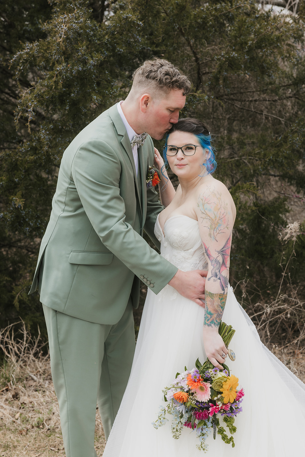 A couple in wedding attire stands outdoors; the groom leans in to kiss the bride, who holds a colorful bouquet. Leafless trees and sparse grass are in the background for a wedding at Discoveryland Ranch
