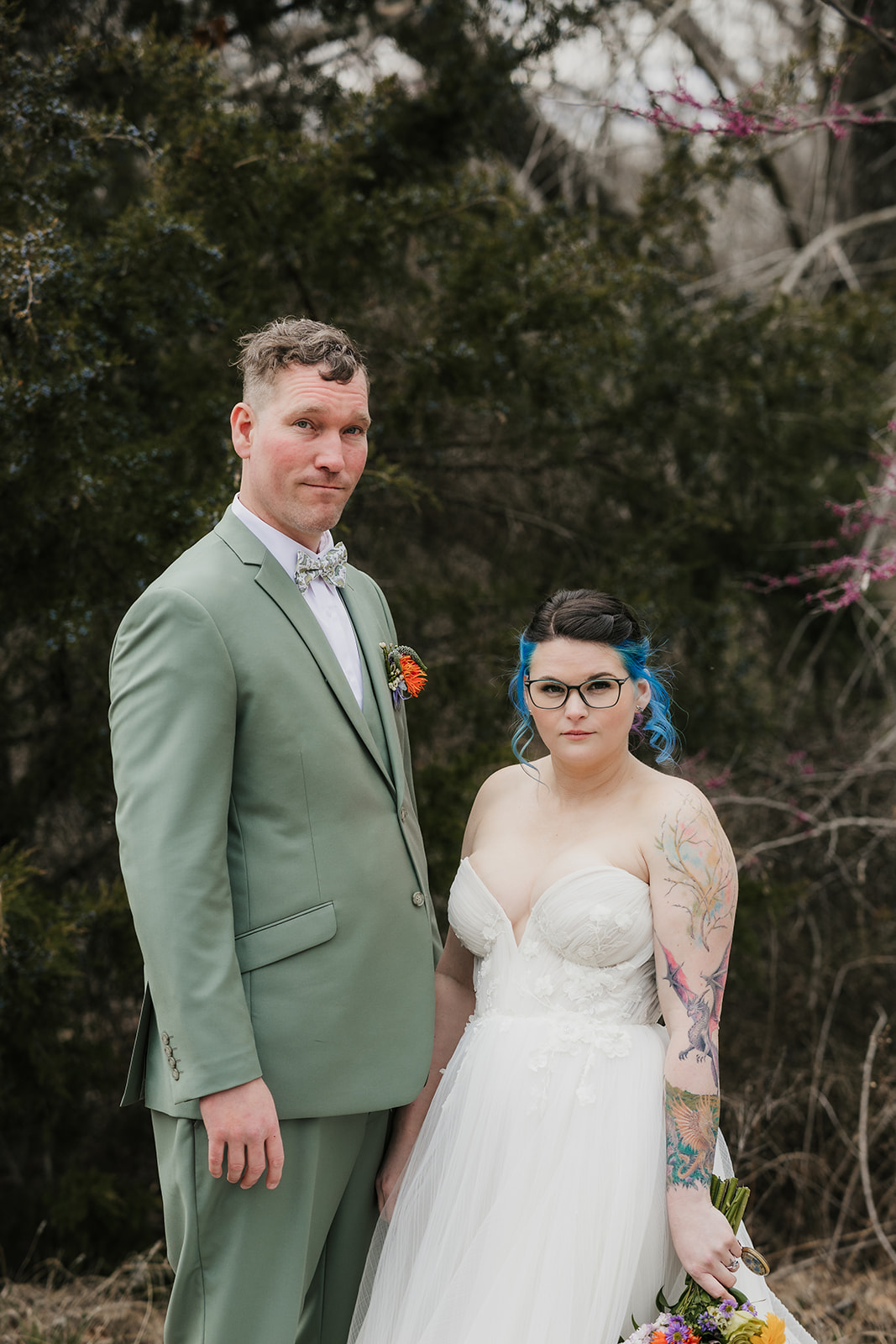 A couple in wedding attire stands outdoors; the groom leans in to kiss the bride, who holds a colorful bouquet. Leafless trees and sparse grass are in the background for a wedding at Discoveryland Ranch