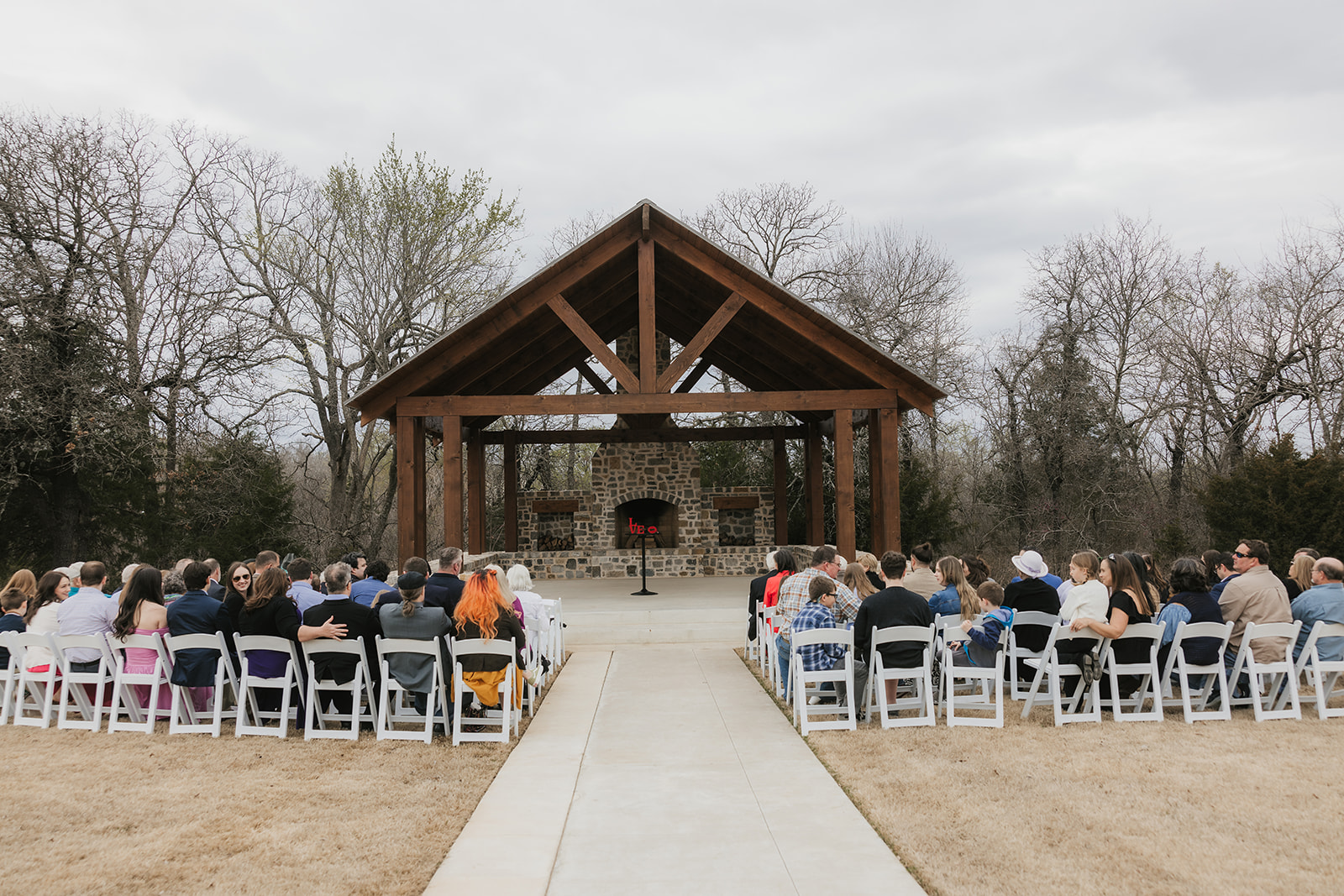 A large group of people seated outdoors facing a wooden pavilion with a stone fireplace, surrounded by leafless trees under a cloudy sky for a wedding at Discoveryland Ranch Wedding