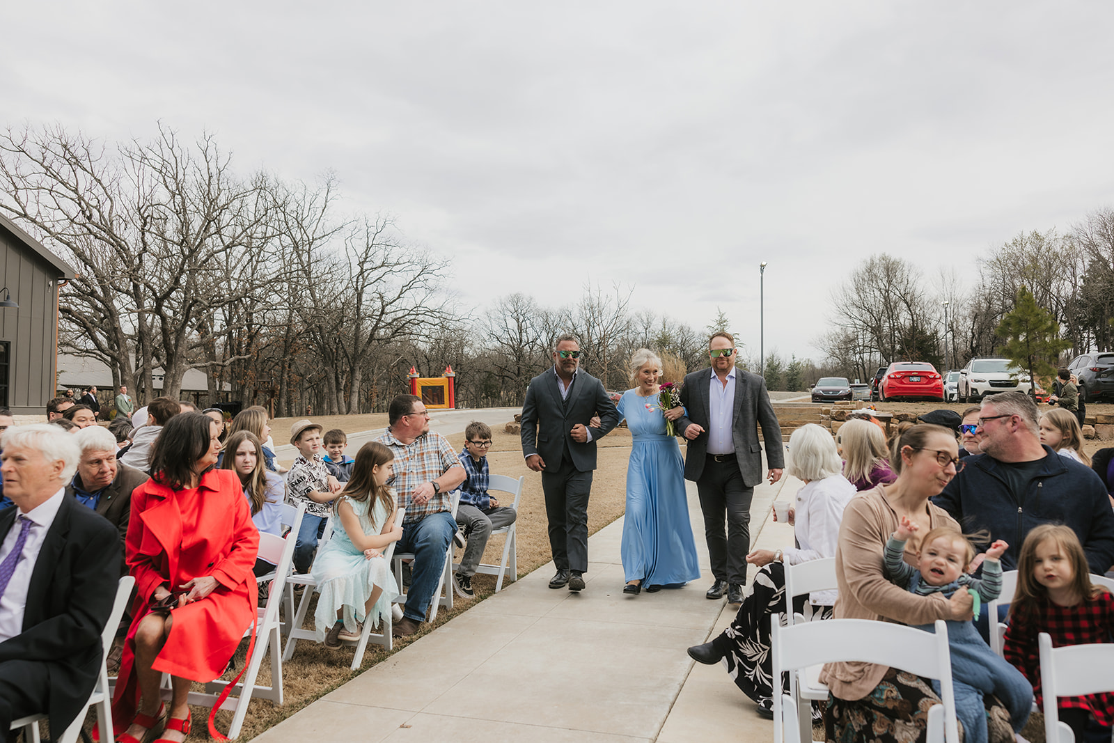A large group of people seated outdoors facing a wooden pavilion with a stone fireplace, surrounded by leafless trees under a cloudy sky for a wedding at Discoveryland Ranch Wedding