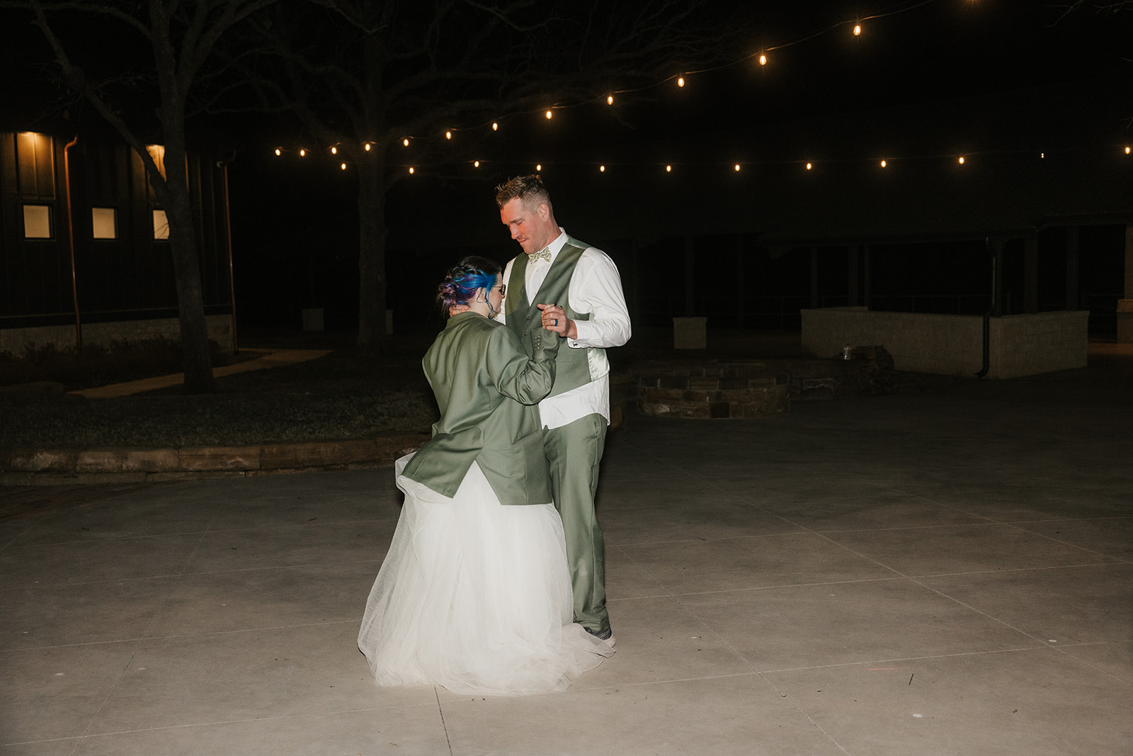 A bride and groom dance outdoors at night under string lights; the bride is sitting while the groom stands, both wearing green jackets.