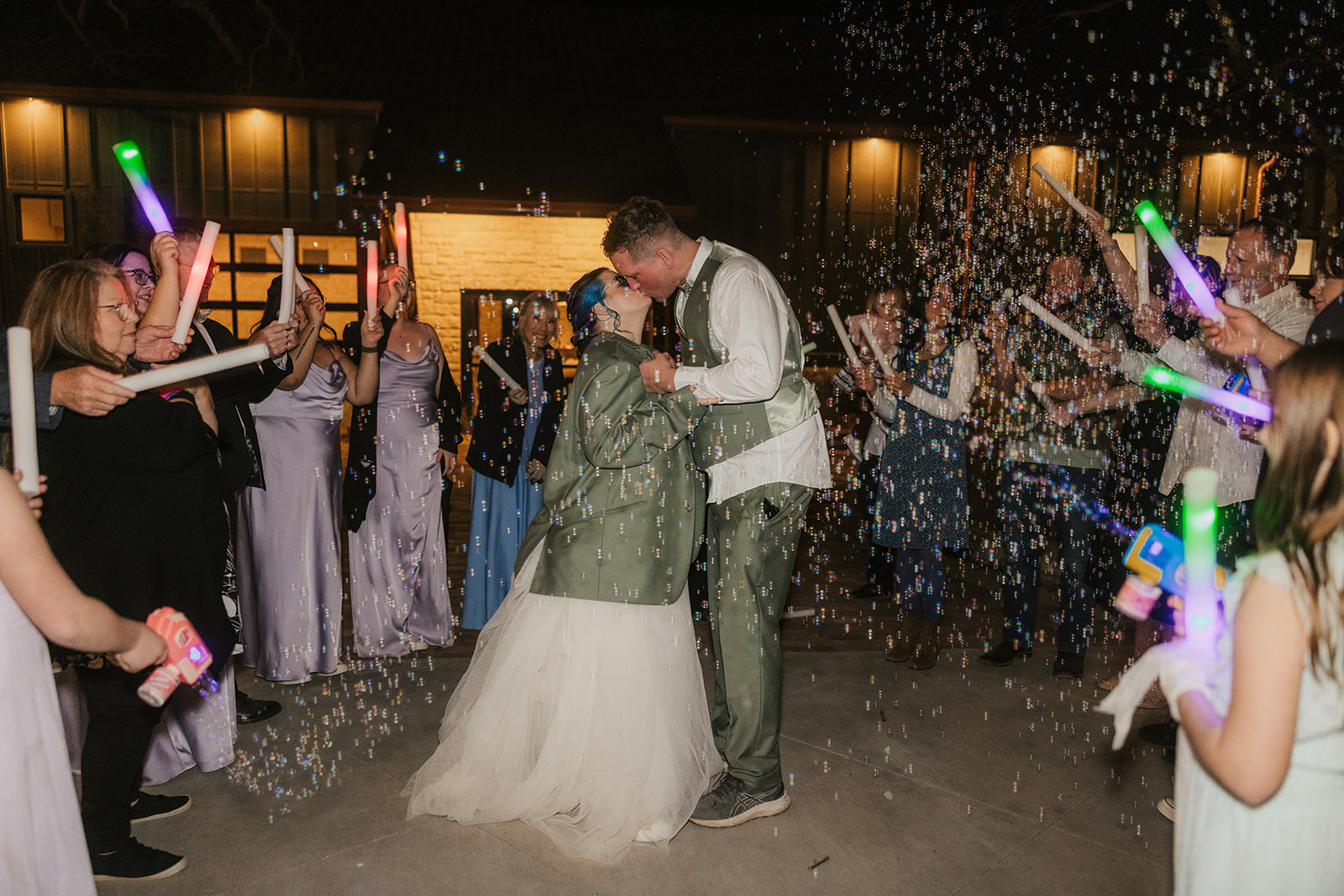 A bride and groom kiss outdoors at night surrounded by guests holding glow sticks and bubbles.