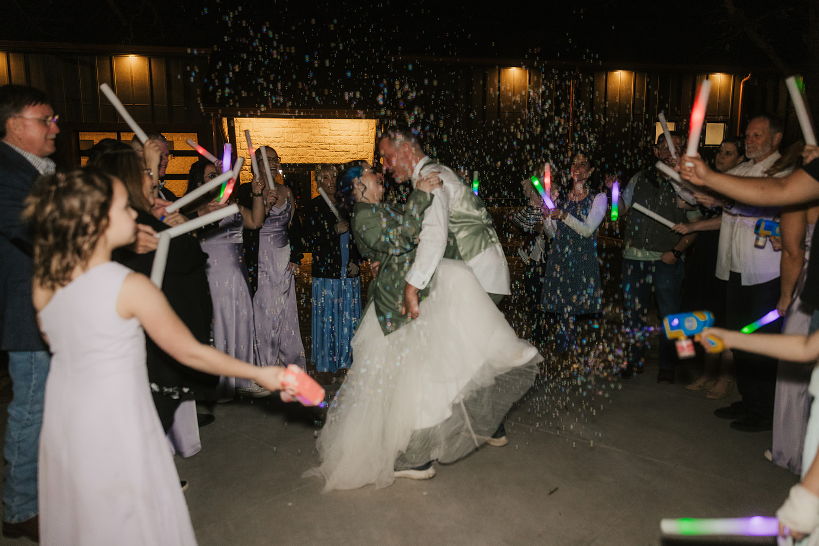 A bride and groom kiss outdoors at night surrounded by guests holding glow sticks and bubbles.