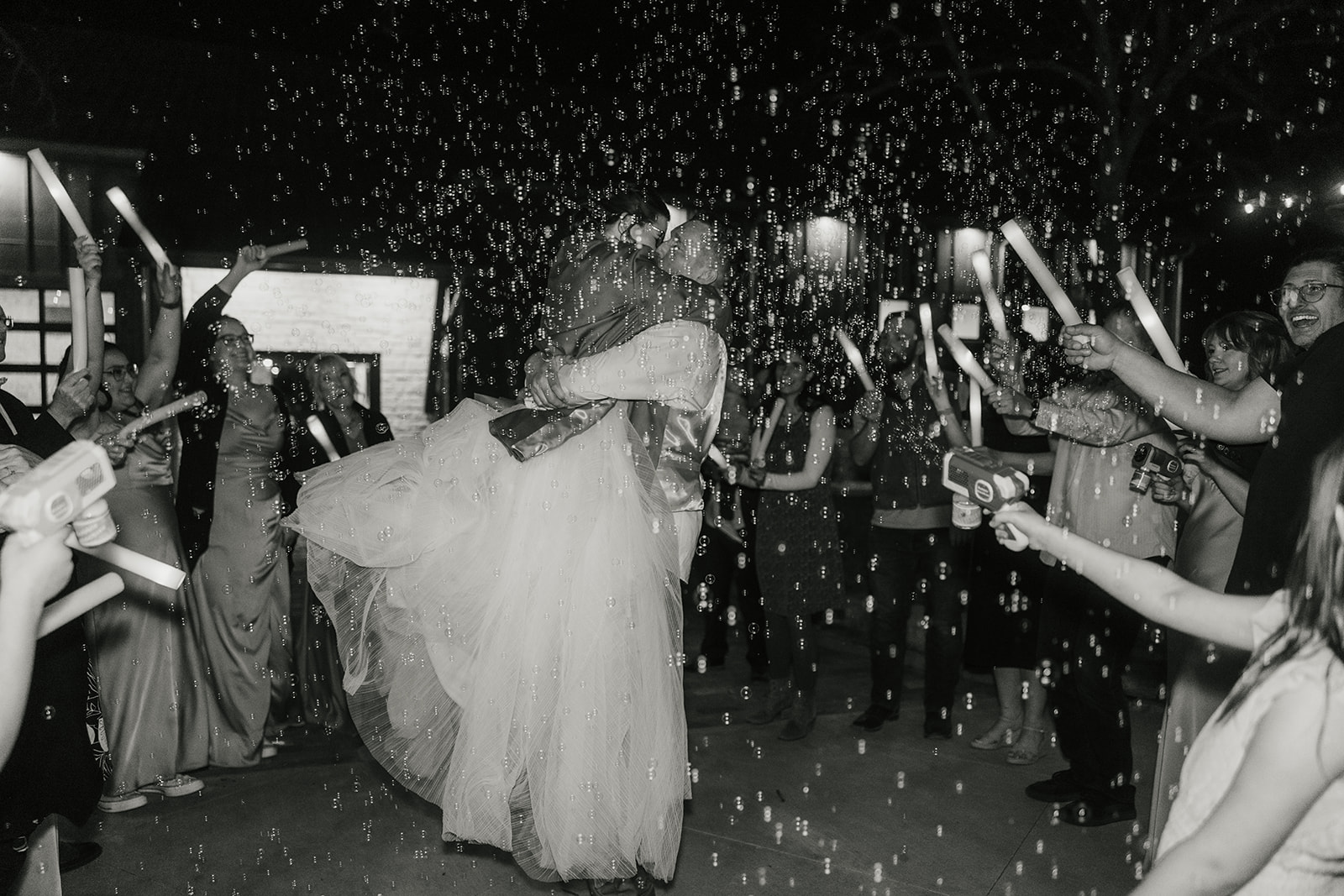 A bride and groom kiss outdoors at night surrounded by guests holding glow sticks and bubbles.