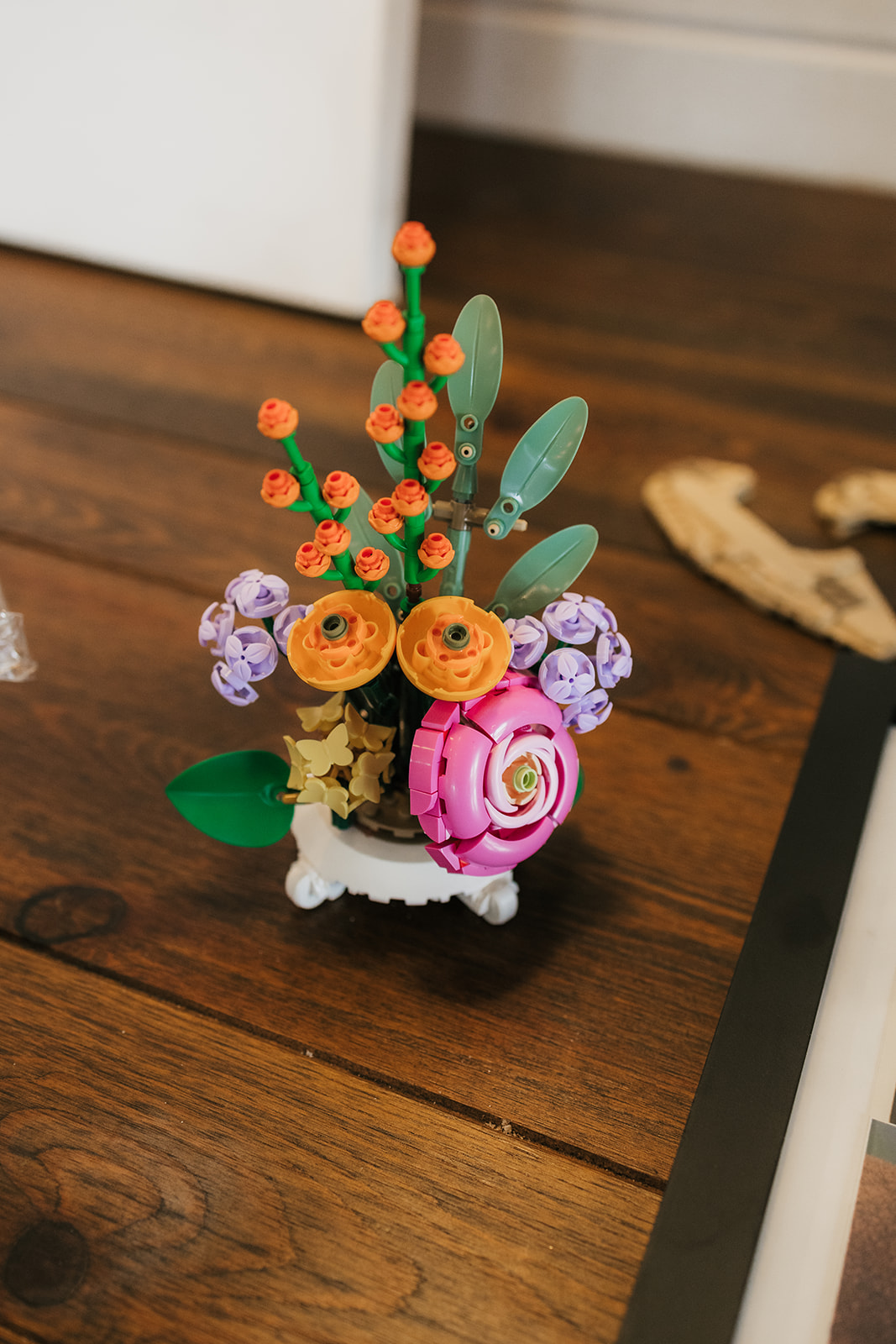 A small decorative flower arrangement with orange, purple, and pink flowers in a white pot sits on a wooden floor.