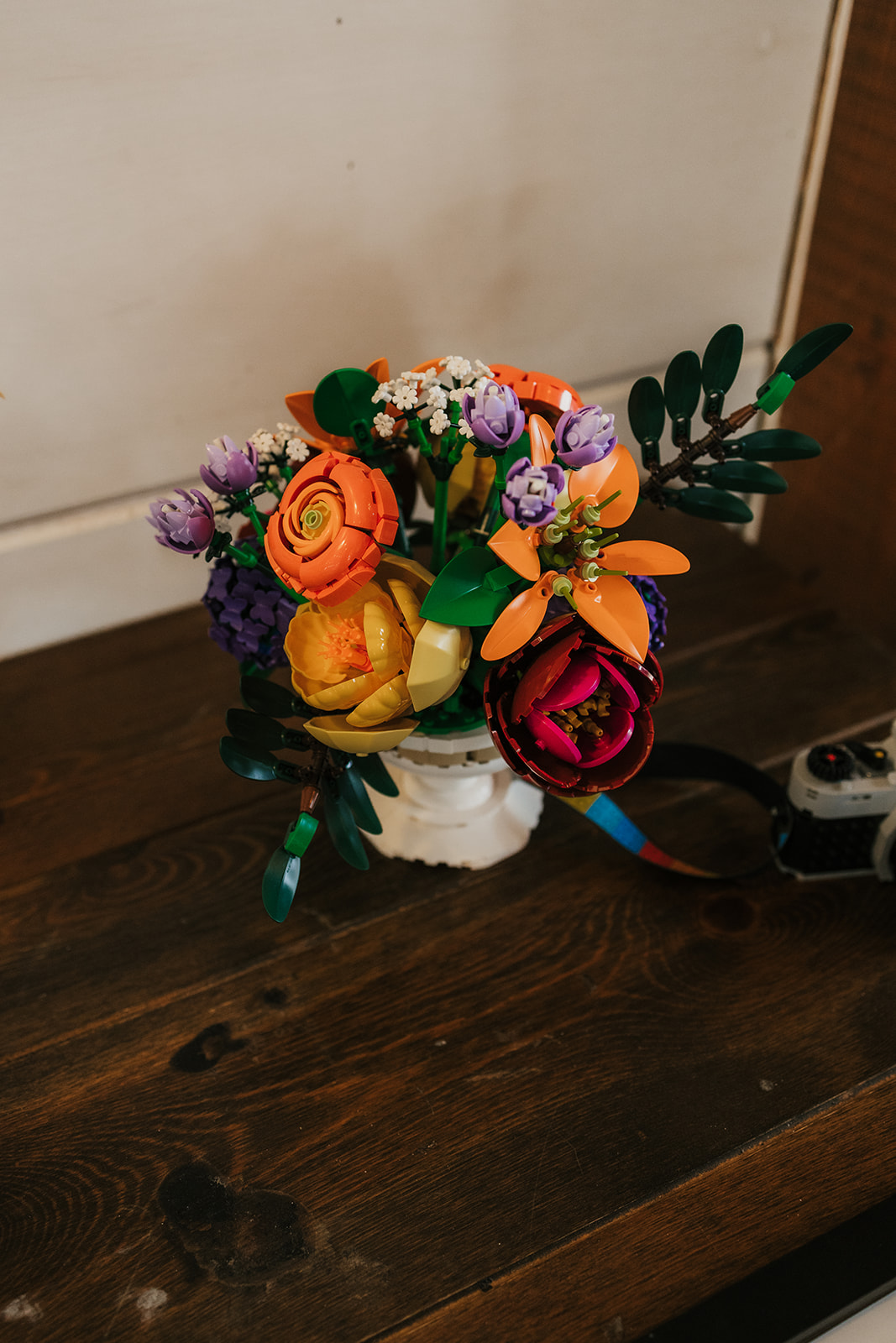 A small decorative flower arrangement with orange, purple, and pink flowers in a white pot sits on a wooden floor.