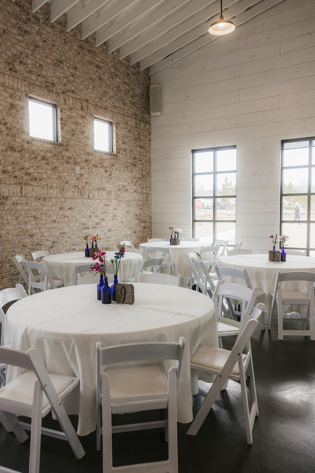 A small event space with round tables covered in white tablecloths, white chairs, and simple floral centerpieces; brick and white walls with several windows.