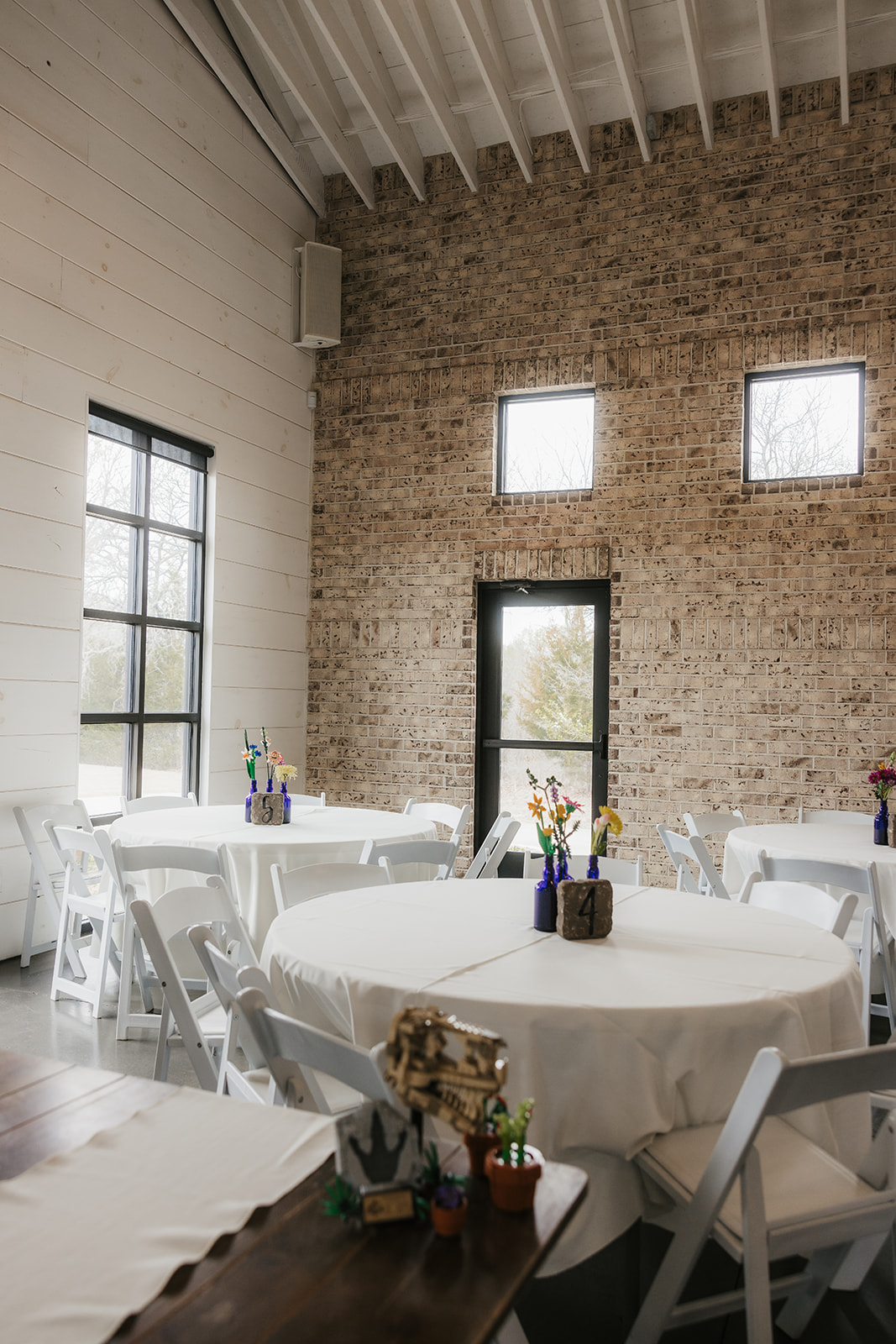 A small event space with round tables covered in white tablecloths, white chairs, and simple floral centerpieces; brick and white walls with several windows.