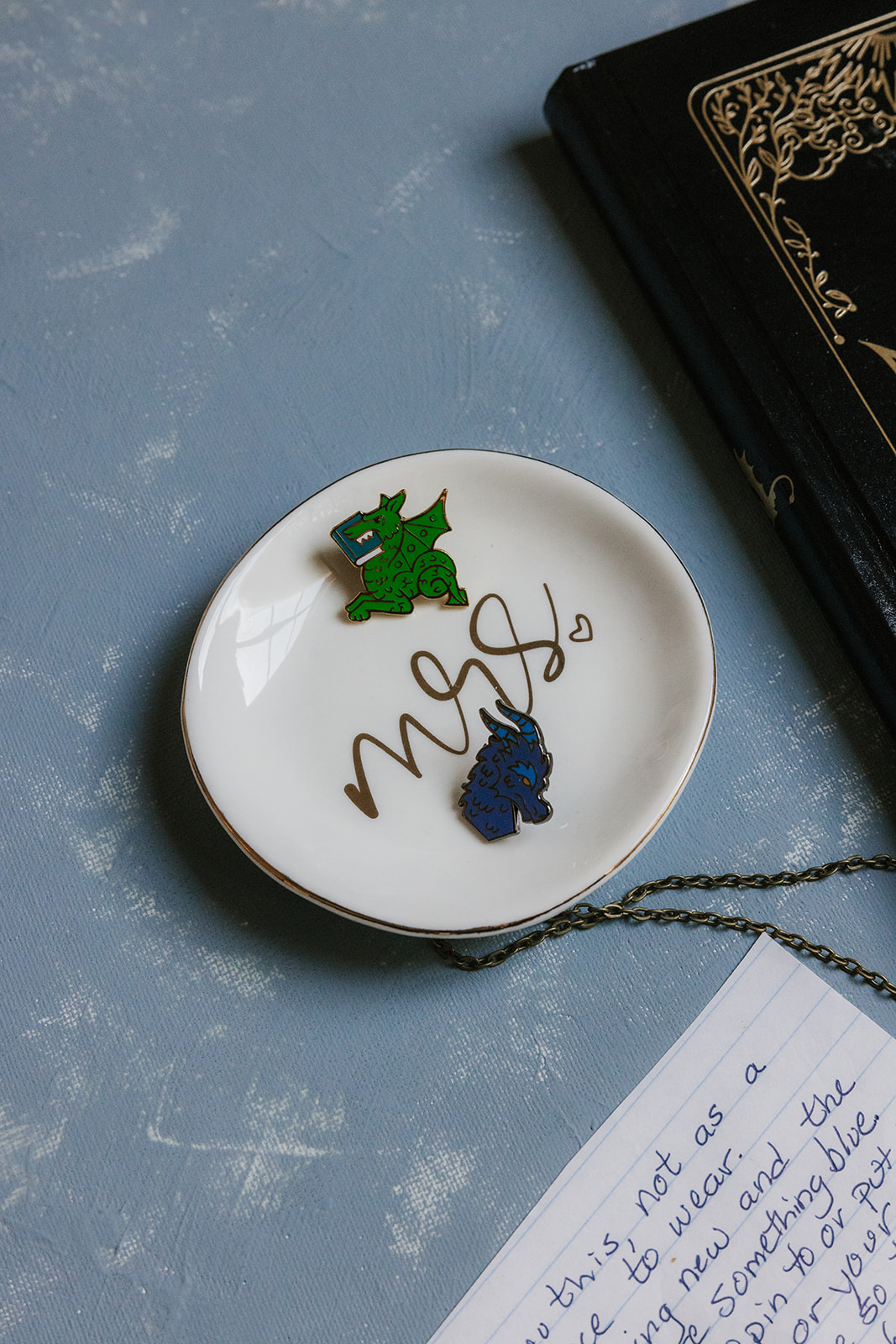 Two enamel pins, one blue and one green, shaped like animals are placed on a silver tray beside yellow wedding invitation cards.