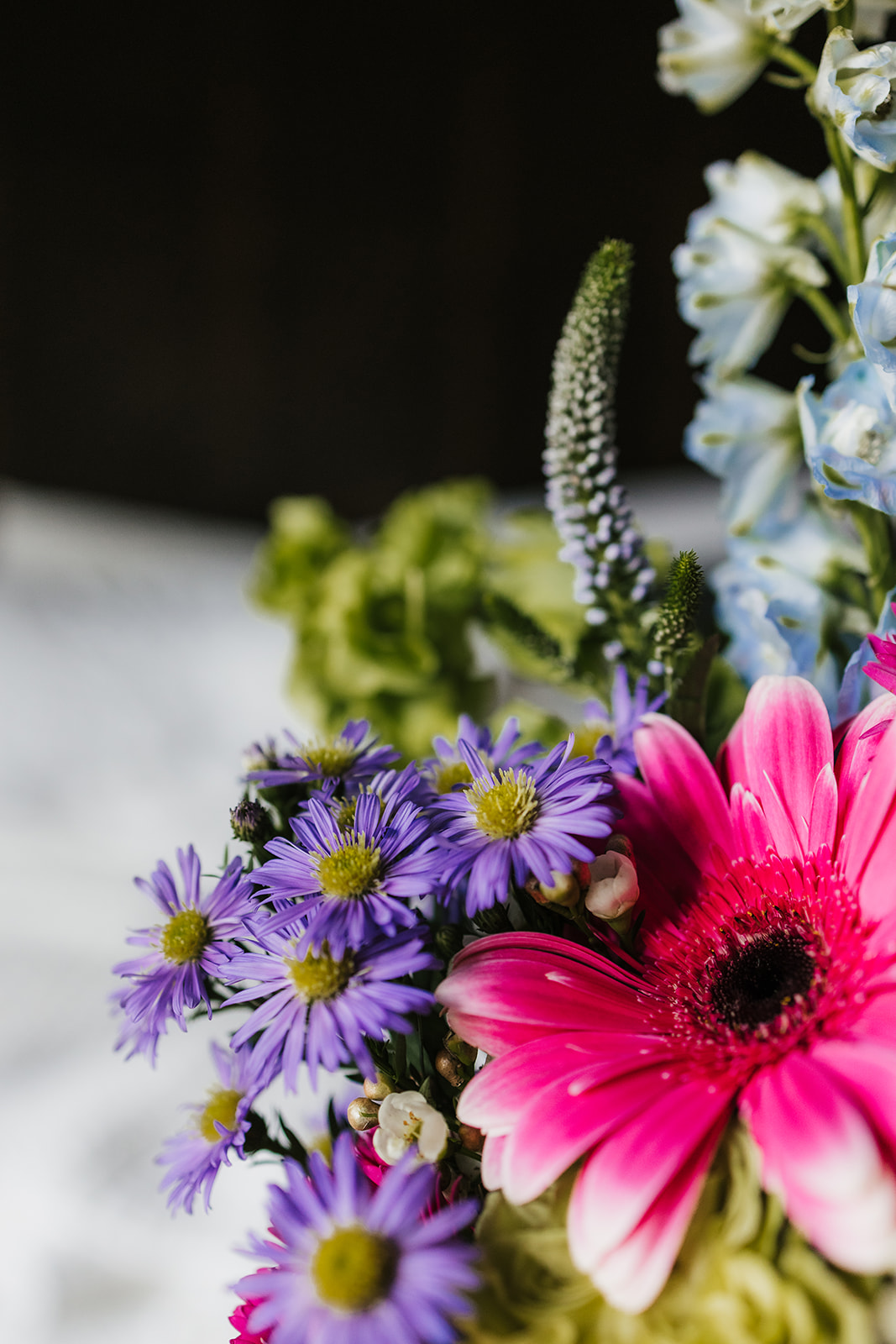 Close-up of a bouquet featuring purple daisies, a bright pink gerbera daisy, and light blue flowers against a dark background.