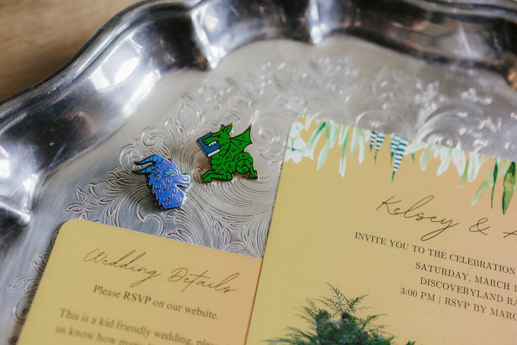 Two enamel pins, one blue and one green, shaped like animals are placed on a silver tray beside yellow wedding invitation cards.