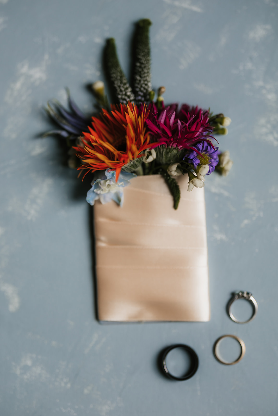 A small bouquet of colorful flowers wrapped in a beige ribbon, with three rings placed on a blue textured surface nearby.