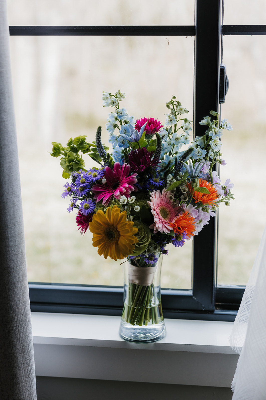 A clear glass vase with a colorful bouquet of assorted flowers sits on a windowsill in front of a black-framed window.