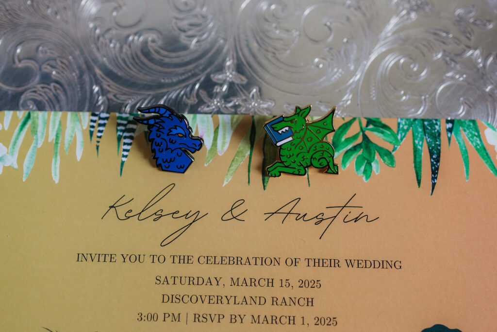 Two enamel pins, one blue and one green, shaped like animals are placed on a silver tray beside yellow wedding invitation cards.