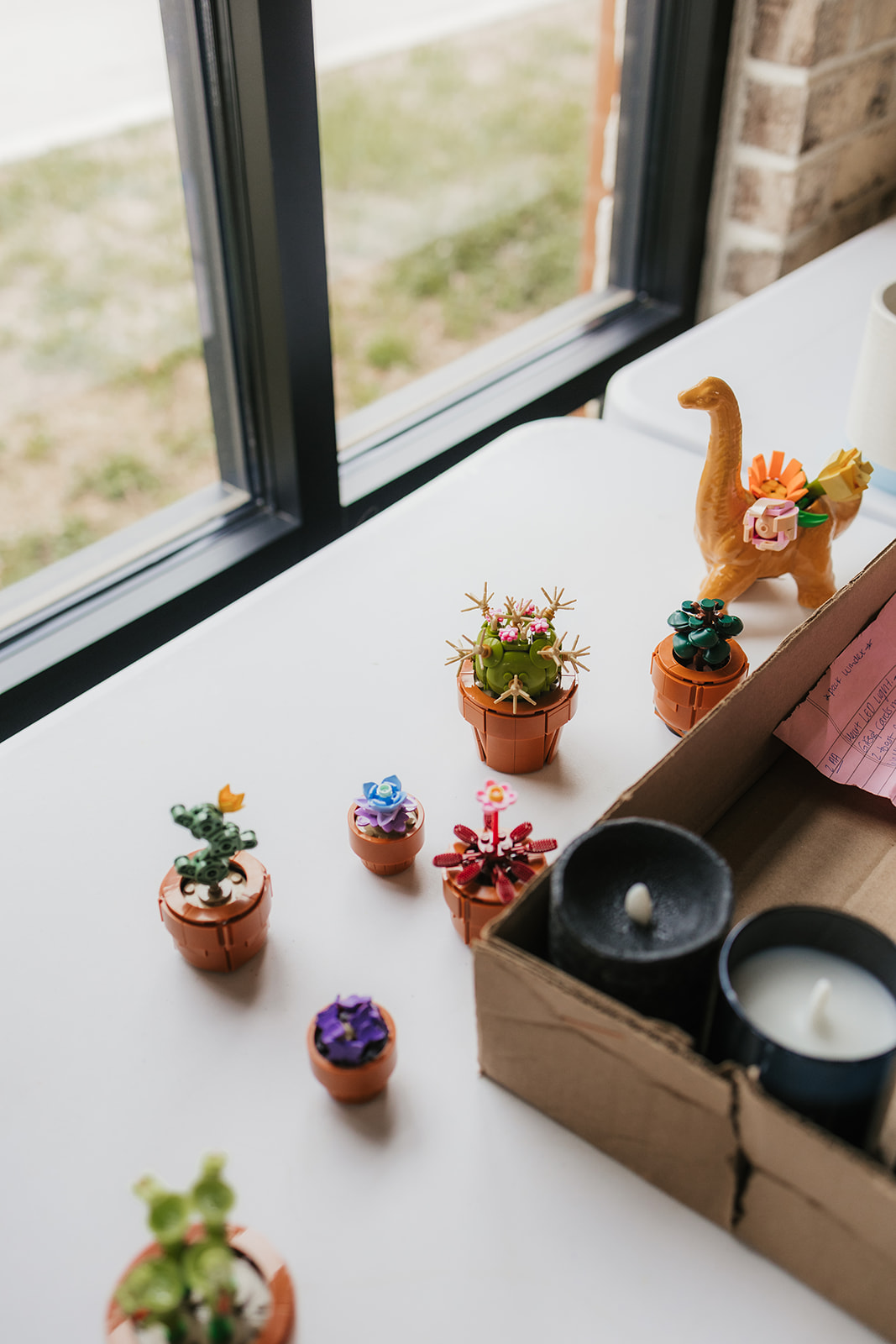 Miniature potted plants and candles are arranged on a white table near a window, with a brown box and a decorative animal planter.