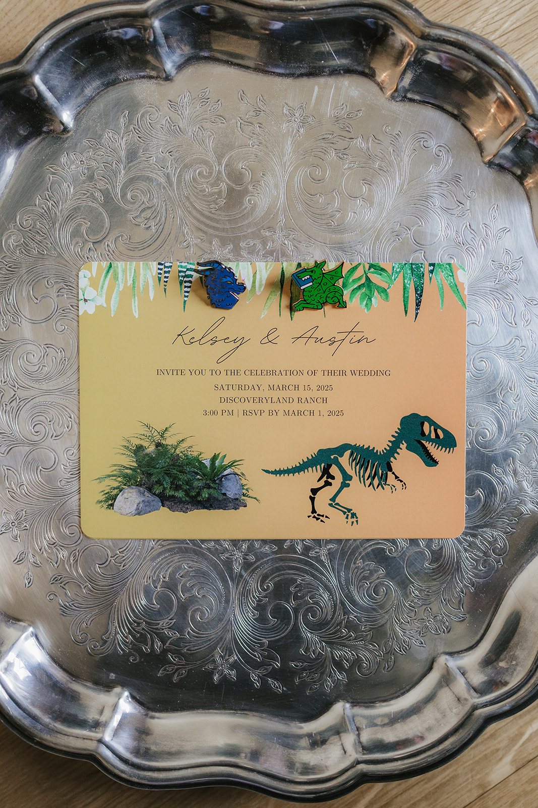 Two enamel pins, one blue and one green, shaped like animals are placed on a silver tray beside yellow wedding invitation cards.