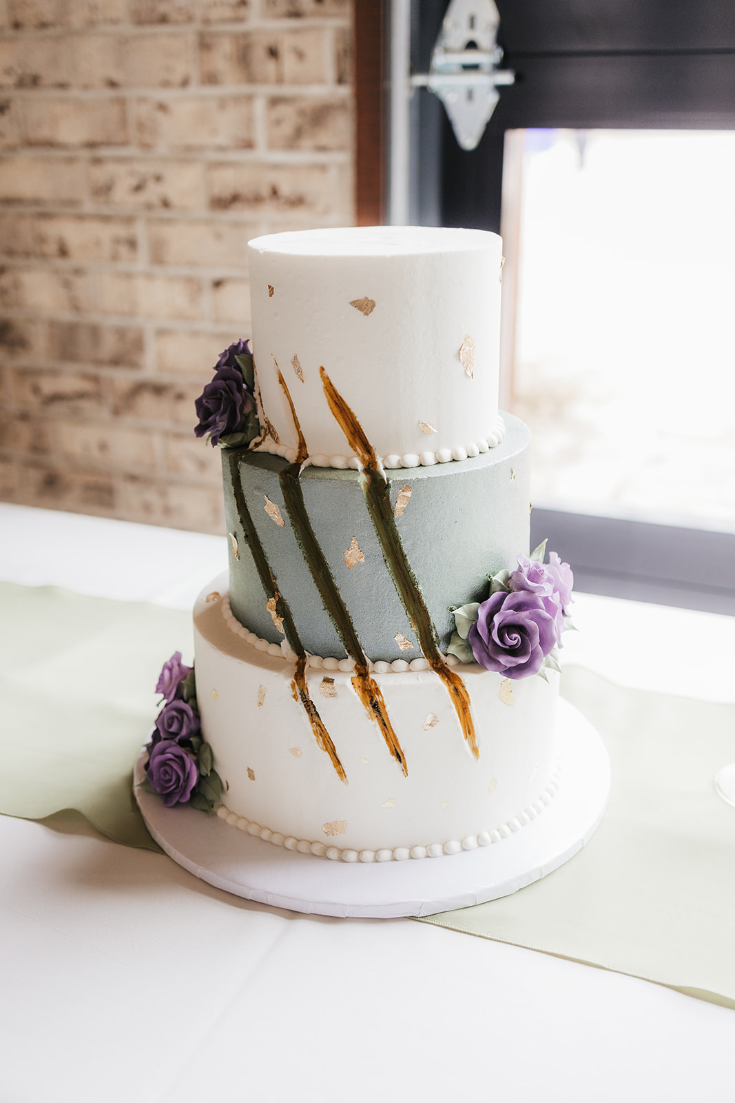 Three-tiered cake with white and green fondant, gold accents, and purple floral decorations, displayed on a white table near a brick wall and window.