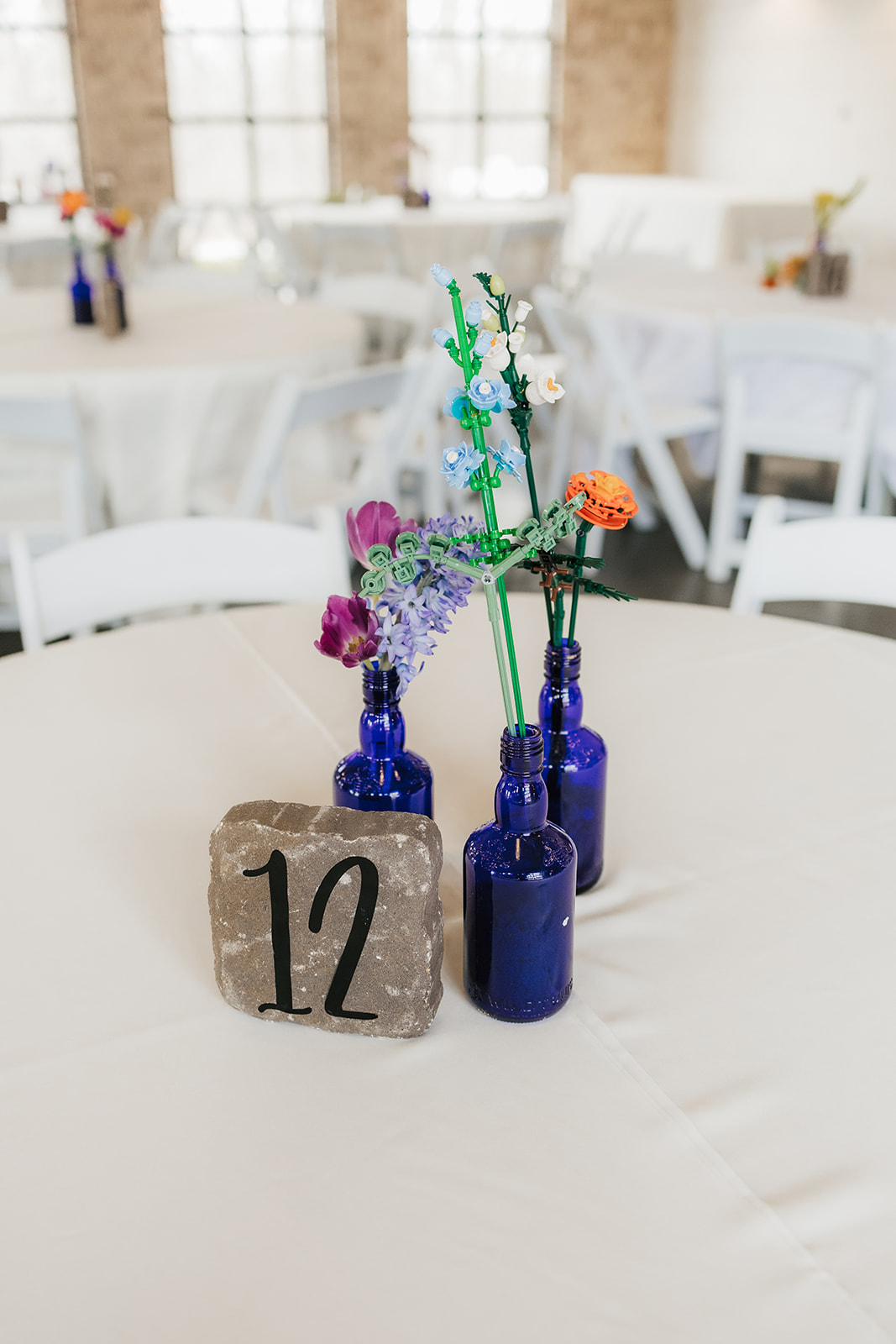 A round table with small blue glass vases holding colorful flowers and a stone displaying the number 12, set in a bright room with white chairs and tables.