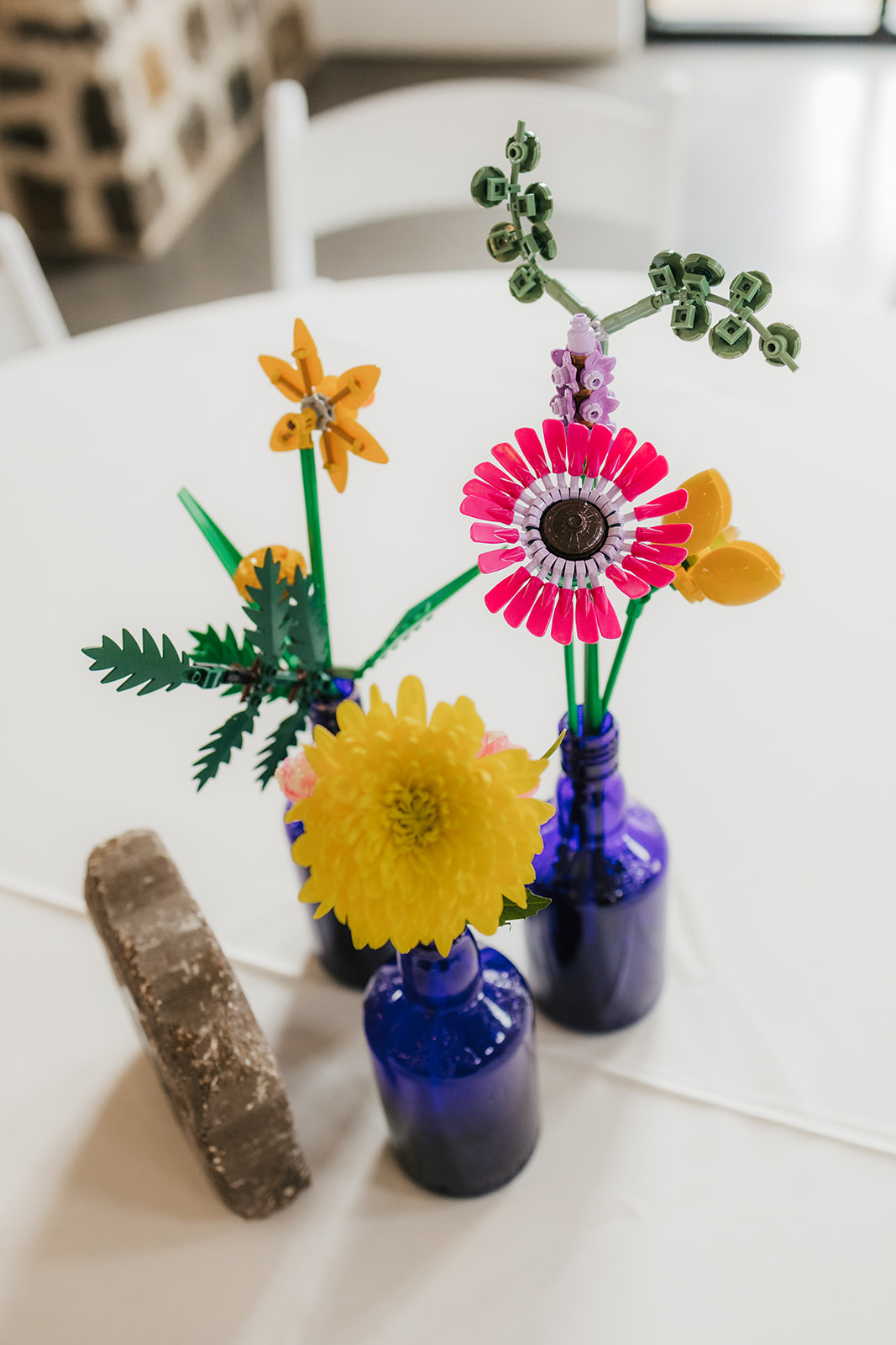 A round table with small blue glass vases holding colorful flowers and a stone displaying the number 12, set in a bright room with white chairs and tables.