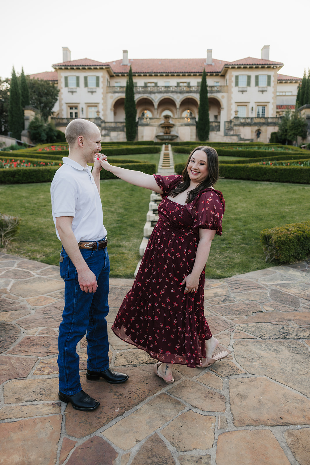 A man and woman hold hands while standing in a manicured garden with red flowers, in front of a large, historic building.