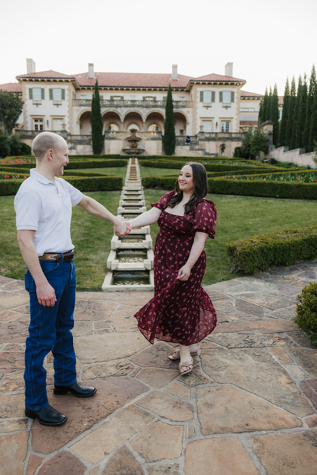 A man and woman hold hands while standing in a manicured garden with red flowers, in front of a large, historic building.