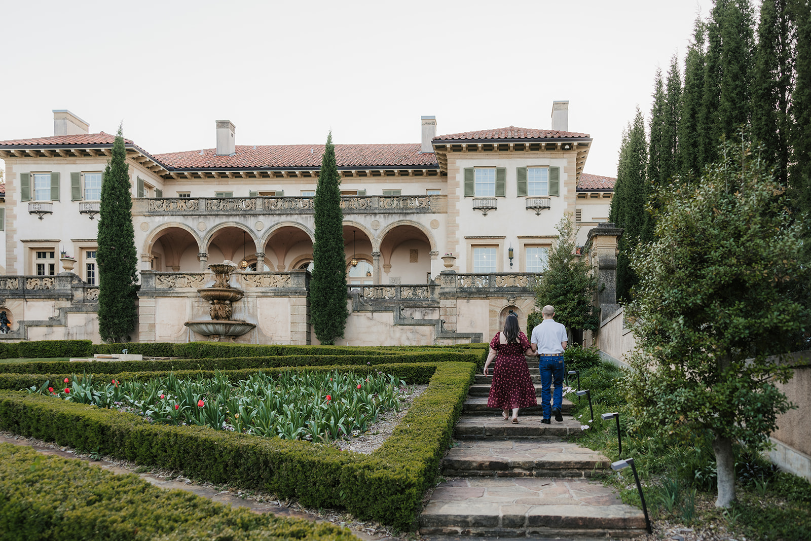 A man and woman hold hands while standing in a manicured garden with red flowers, in front of a large, historic building.