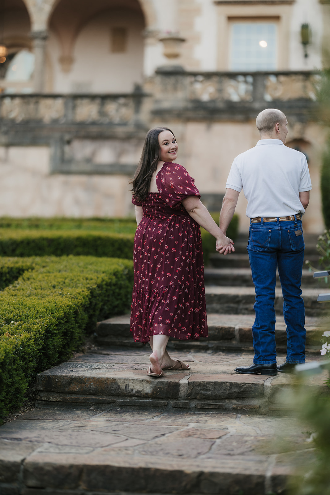 A man and woman hold hands while standing in a manicured garden with red flowers, in front of a large, historic building.