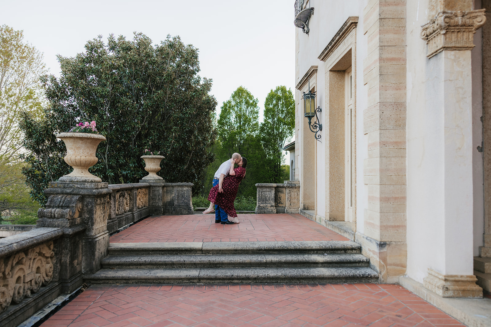 A man and woman hold hands while standing in a manicured garden with red flowers, in front of a large, historic building.
