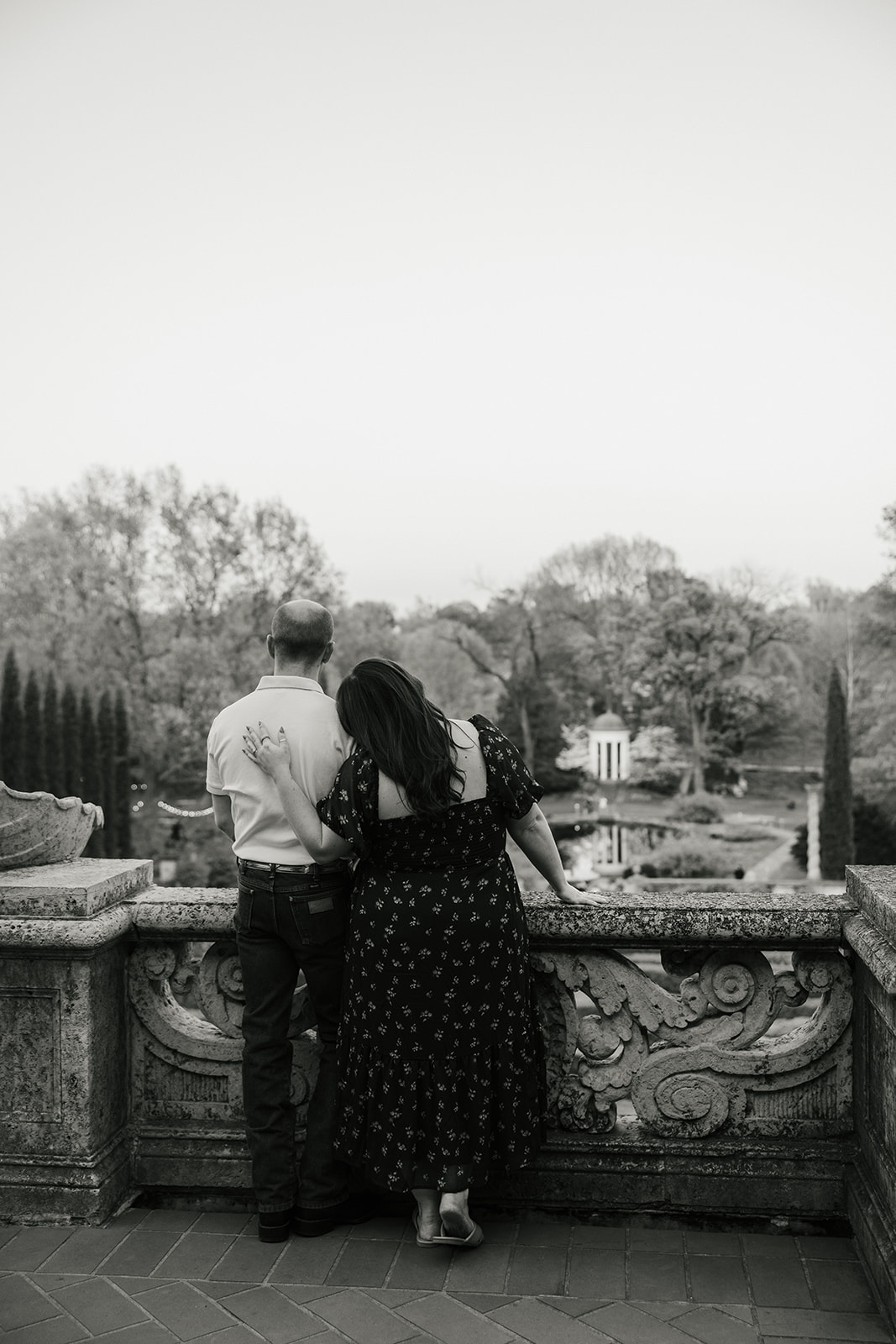 A man and woman hold hands while standing in a manicured garden with red flowers, in front of a large, historic building.