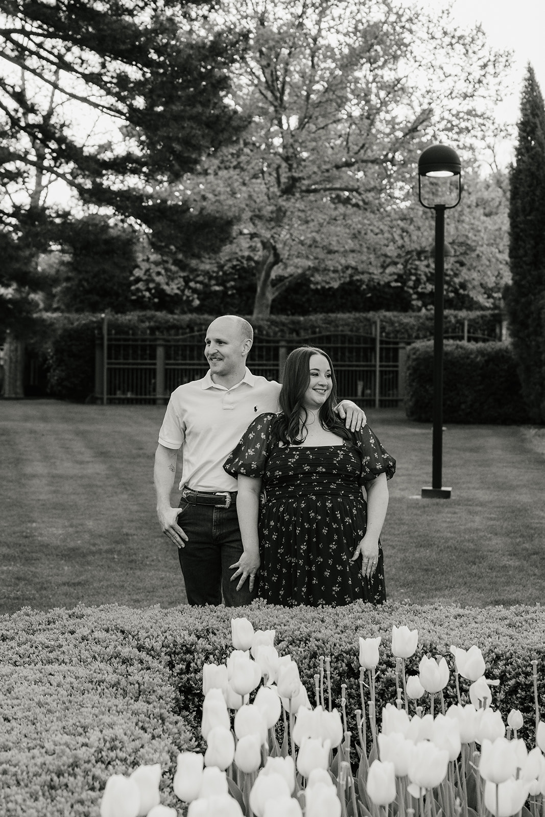 A man and woman hold hands while standing in a manicured garden with red flowers, in front of a large, historic building.
