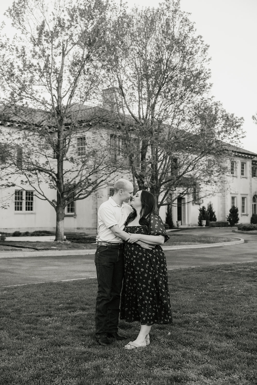 A man and woman hold hands while standing in a manicured garden with red flowers, in front of a large, historic building.