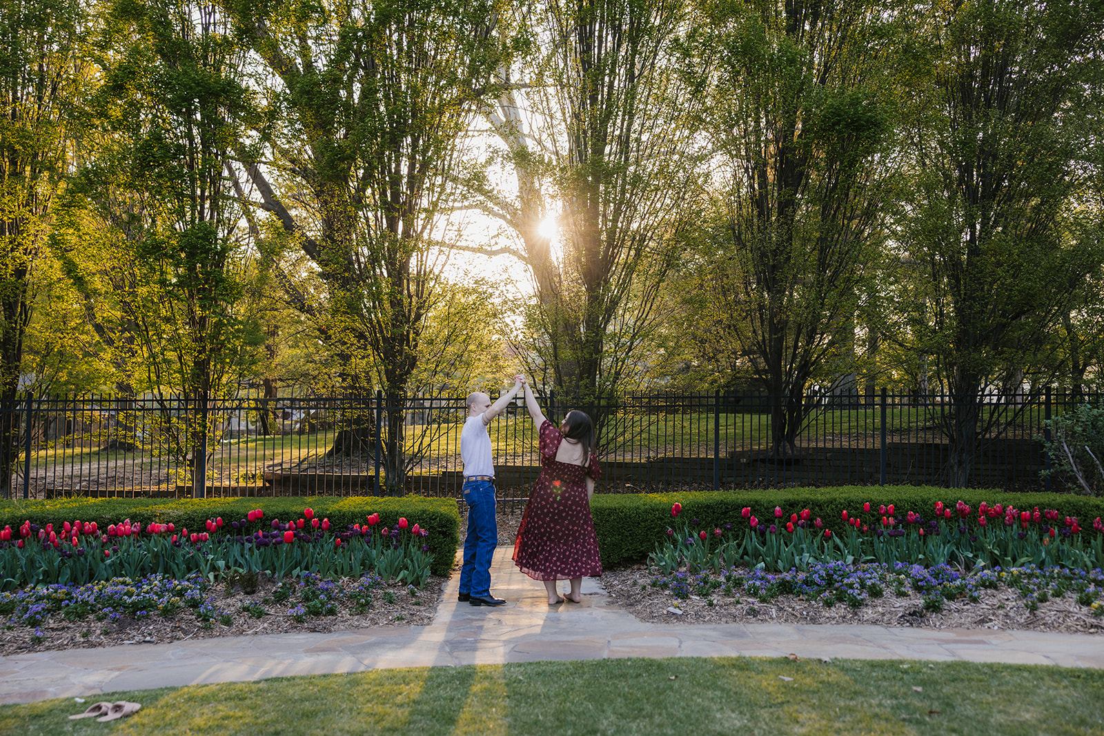 A man and woman dance together on a garden pathway surrounded by tulips and trees, with sunlight shining through the branches.