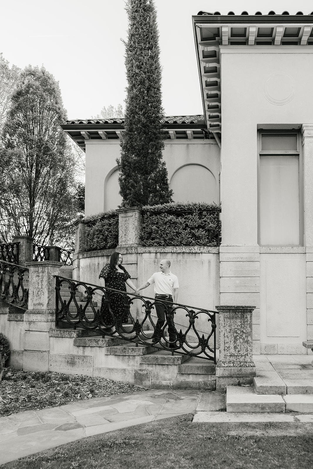 Two people walk up stairs beside a large building with tall windows and decorative railing, surrounded by trees and hedges.