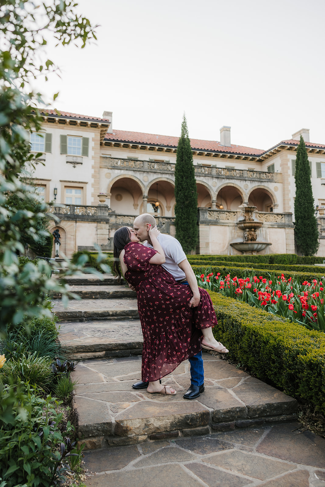 A couple holds hands while walking through a manicured garden with red flowers, in front of a historic building with arches and tall trees.