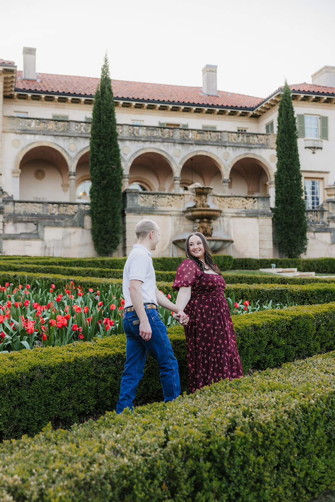 A couple holds hands while walking through a manicured garden with red flowers, in front of a historic building with arches and tall trees.
