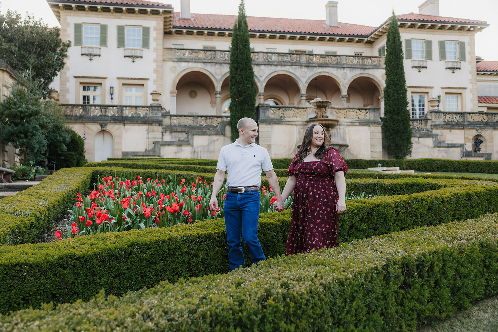 A man and woman hold hands while standing in a manicured garden with red flowers, in front of a large, historic building.