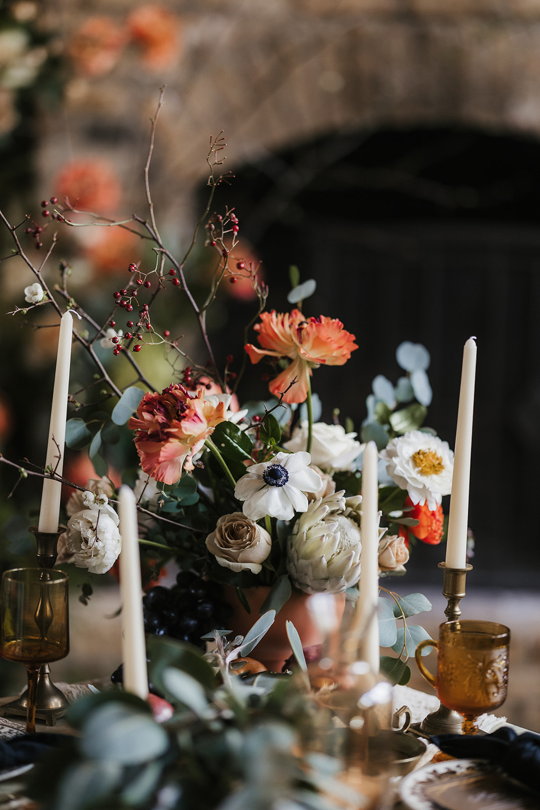 A floral centerpiece with white, pink, and orange flowers, surrounded by tall ivory candles and amber glassware on a table.