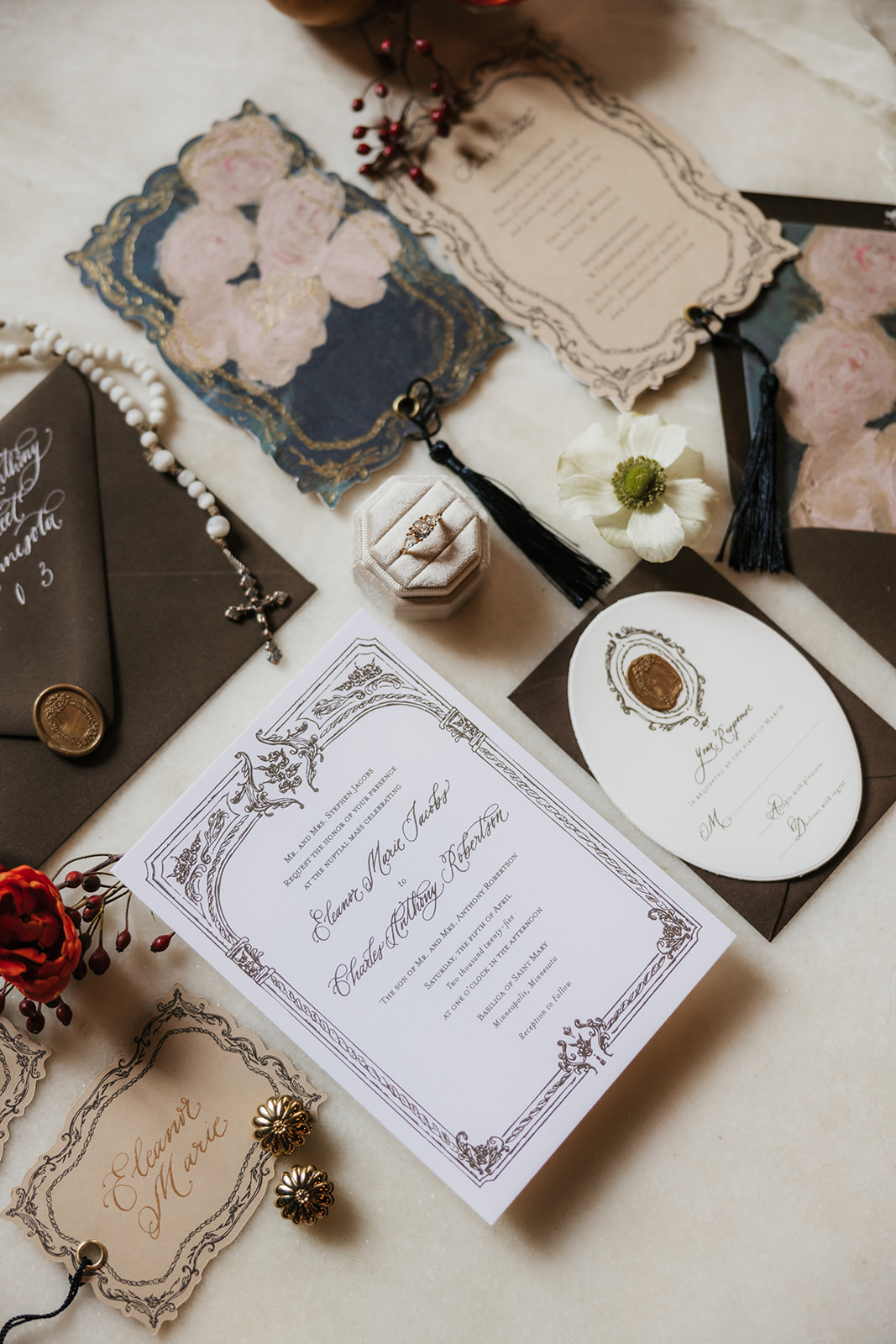 A flat lay of elegant wedding invitations, envelopes, a rosary, a wax seal, and decorative cards arranged on a light surface.