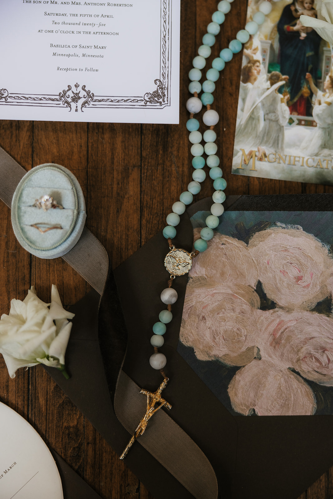 Flat lay of wedding invitations, floral arrangements, a beaded necklace, a photo, and decorative items arranged on a wooden floor for catholic wedding ideas