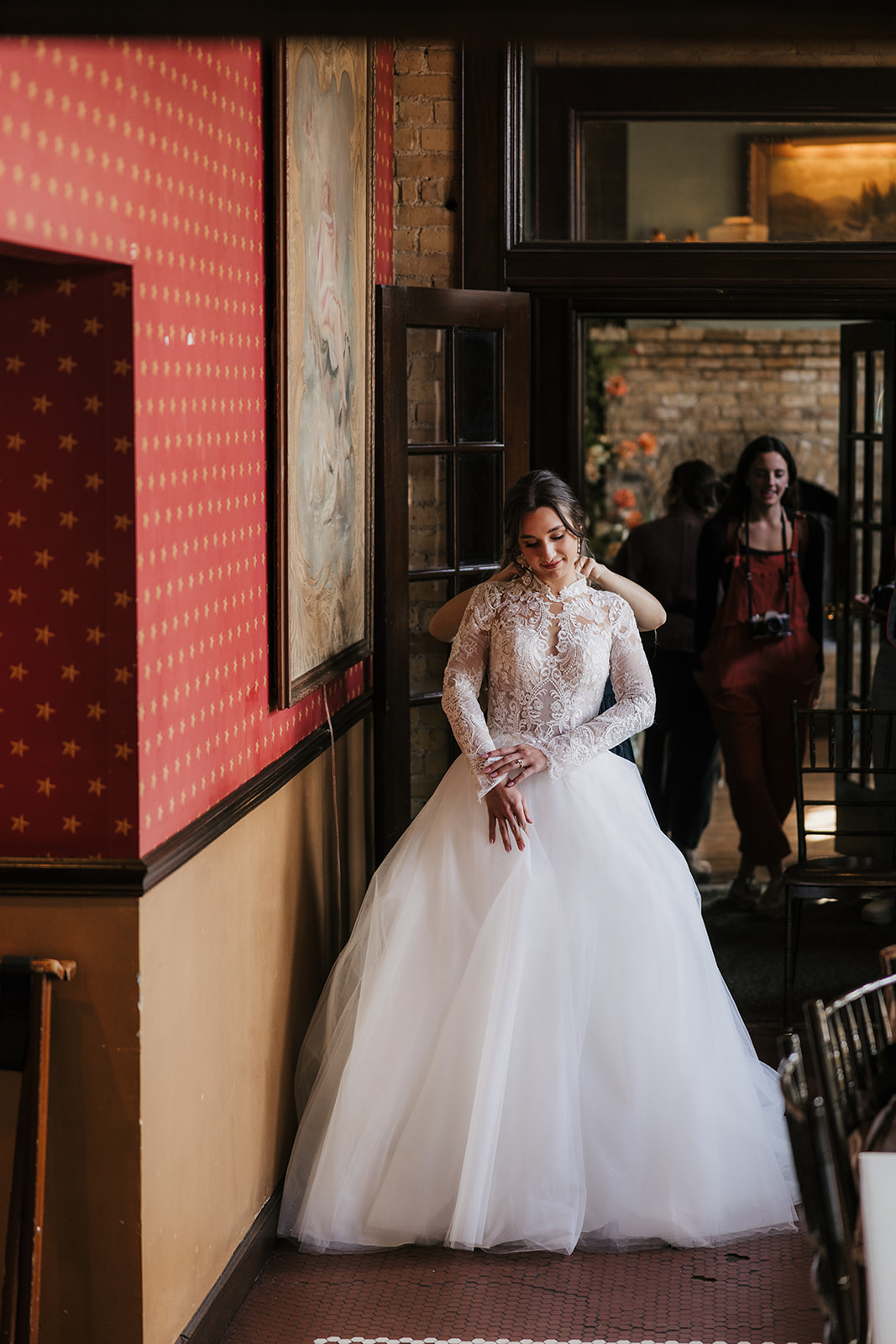 A woman in a white wedding dress stands indoors near a glass door, smiling, with a large classical painting on the wall beside her for catholic wedding ideas
