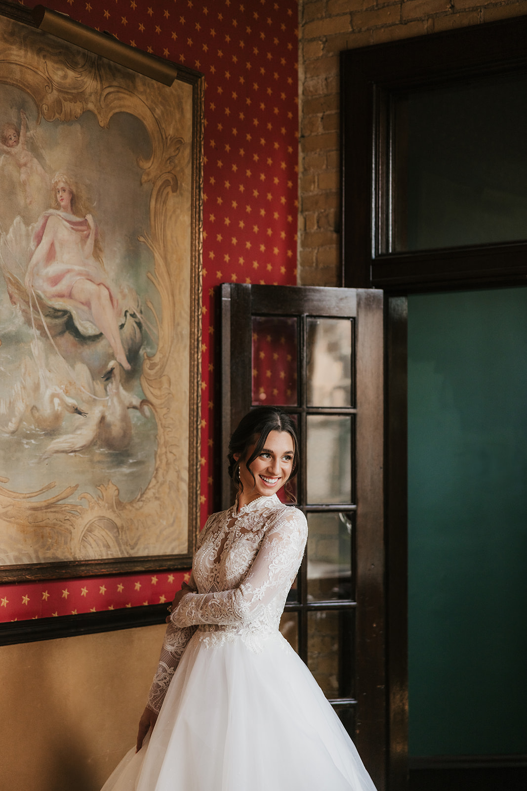 A woman in a white wedding dress stands indoors near a glass door, smiling, with a large classical painting on the wall beside her for catholic wedding ideas