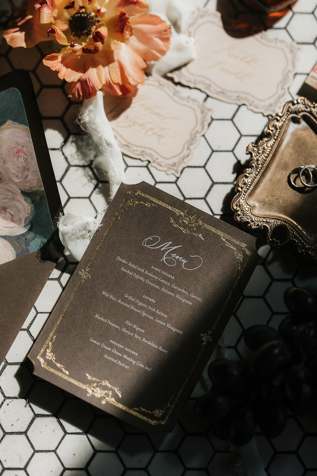 A wedding menu card with gold detailing lies on a tiled surface, surrounded by flowers, ornate cards, a framed mirror, and two rings.