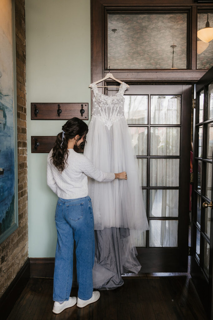 A person in jeans and a white shirt holds a white wedding dress hanging on a door inside a room with wooden floors and glass panels.