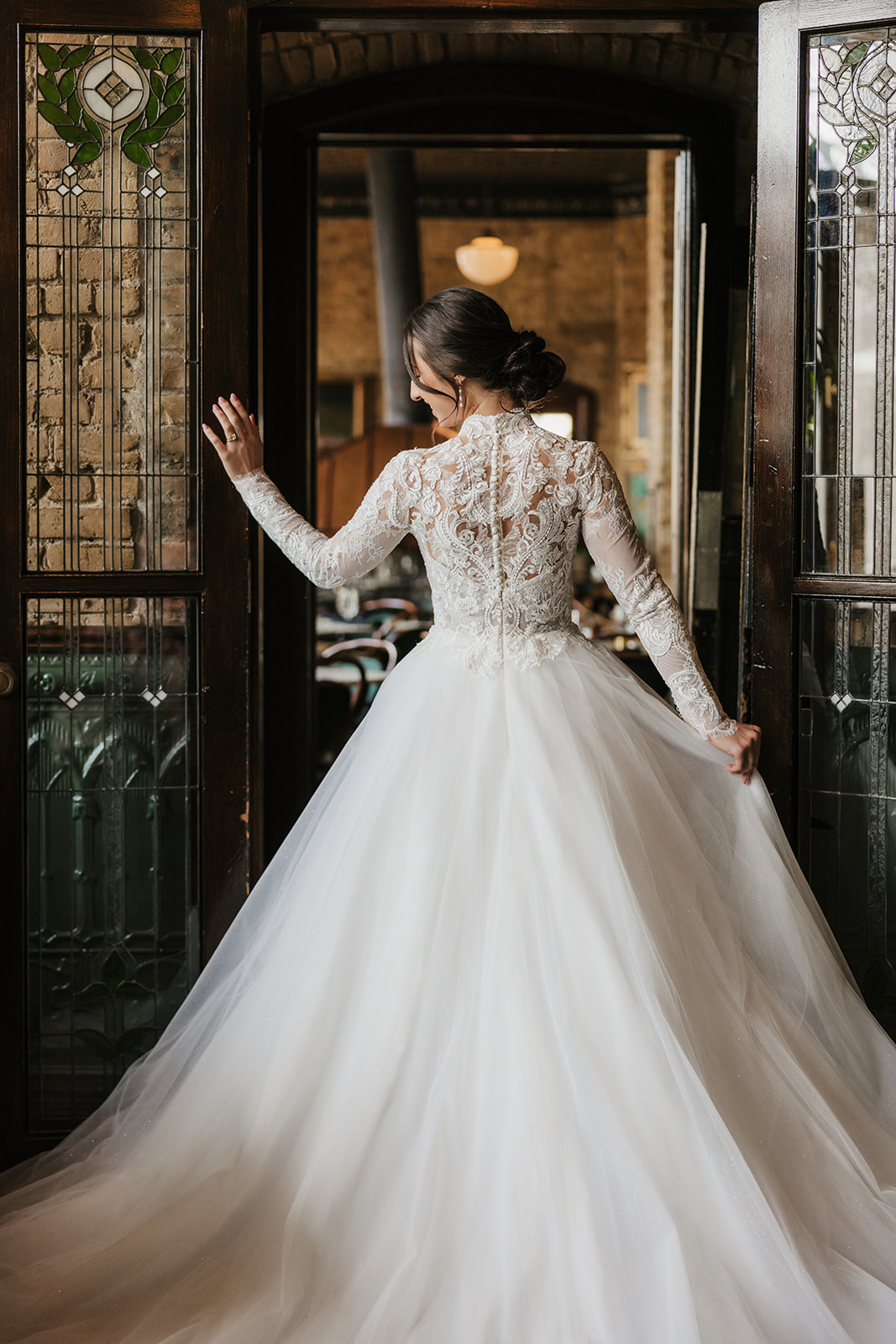 A woman in a white wedding dress stands indoors near a glass door, smiling, with a large classical painting on the wall beside her for catholic wedding ideas