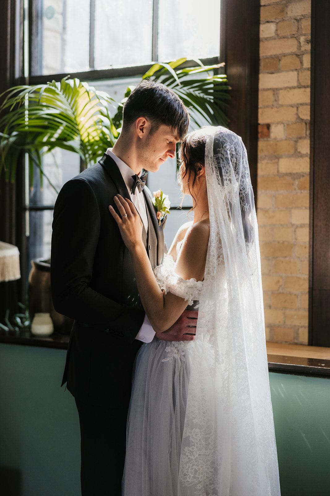 A bride and groom stand close together, gazing at each other in front of a window with sunlight streaming in, surrounded by greenery and brick walls for catholic wedding ideas