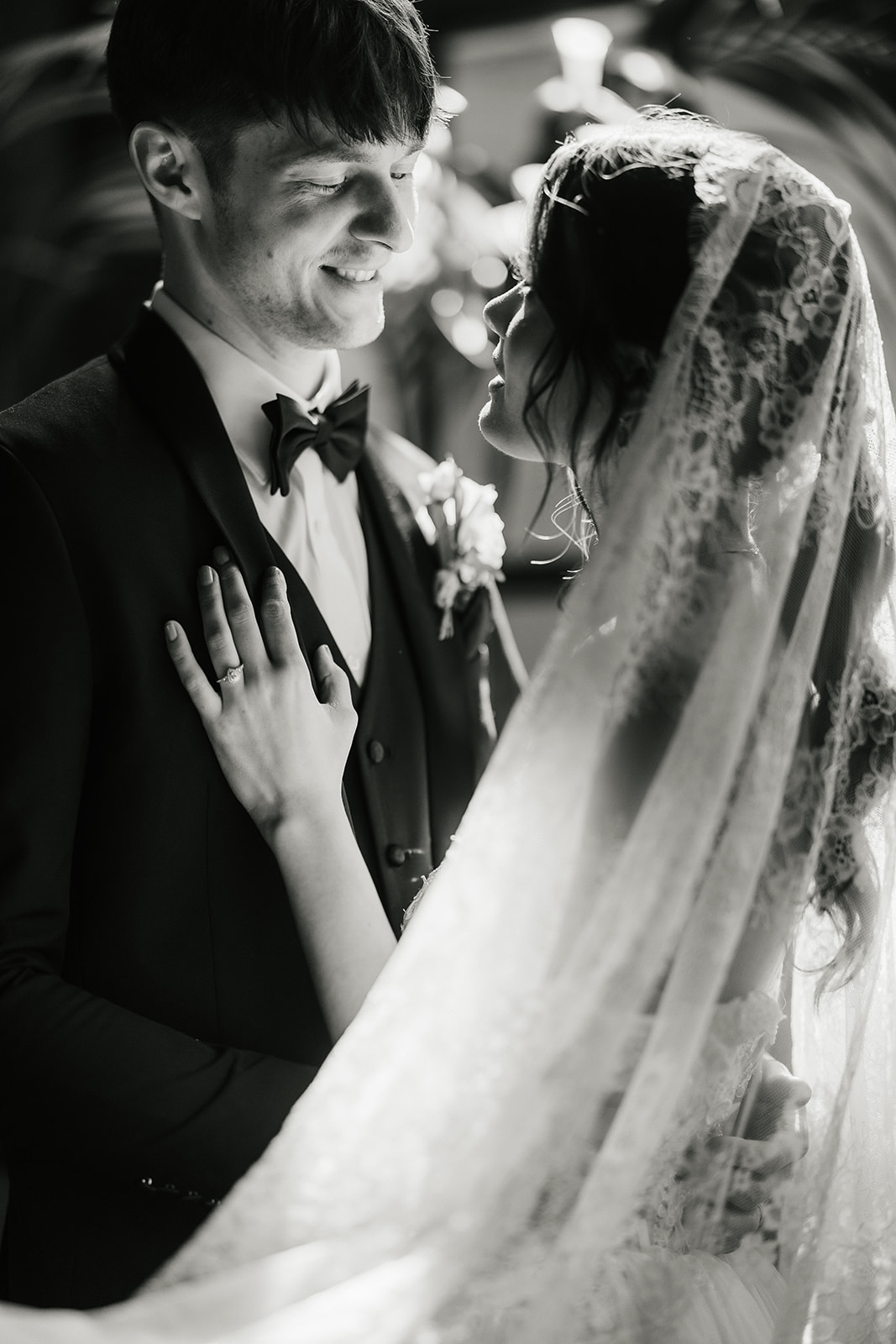 A bride and groom stand close together, gazing at each other in front of a window with sunlight streaming in, surrounded by greenery and brick walls for catholic wedding ideas