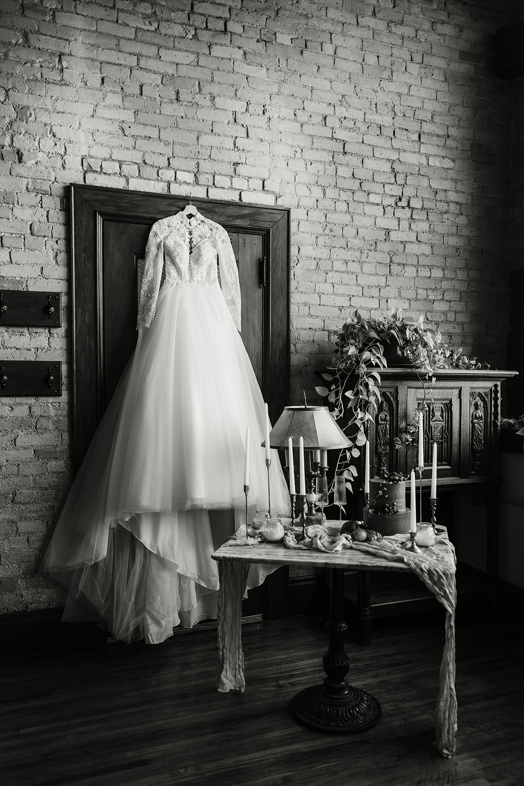 A long-sleeved wedding dress hangs on a door next to a table set with candles and decor in a room with brick walls.