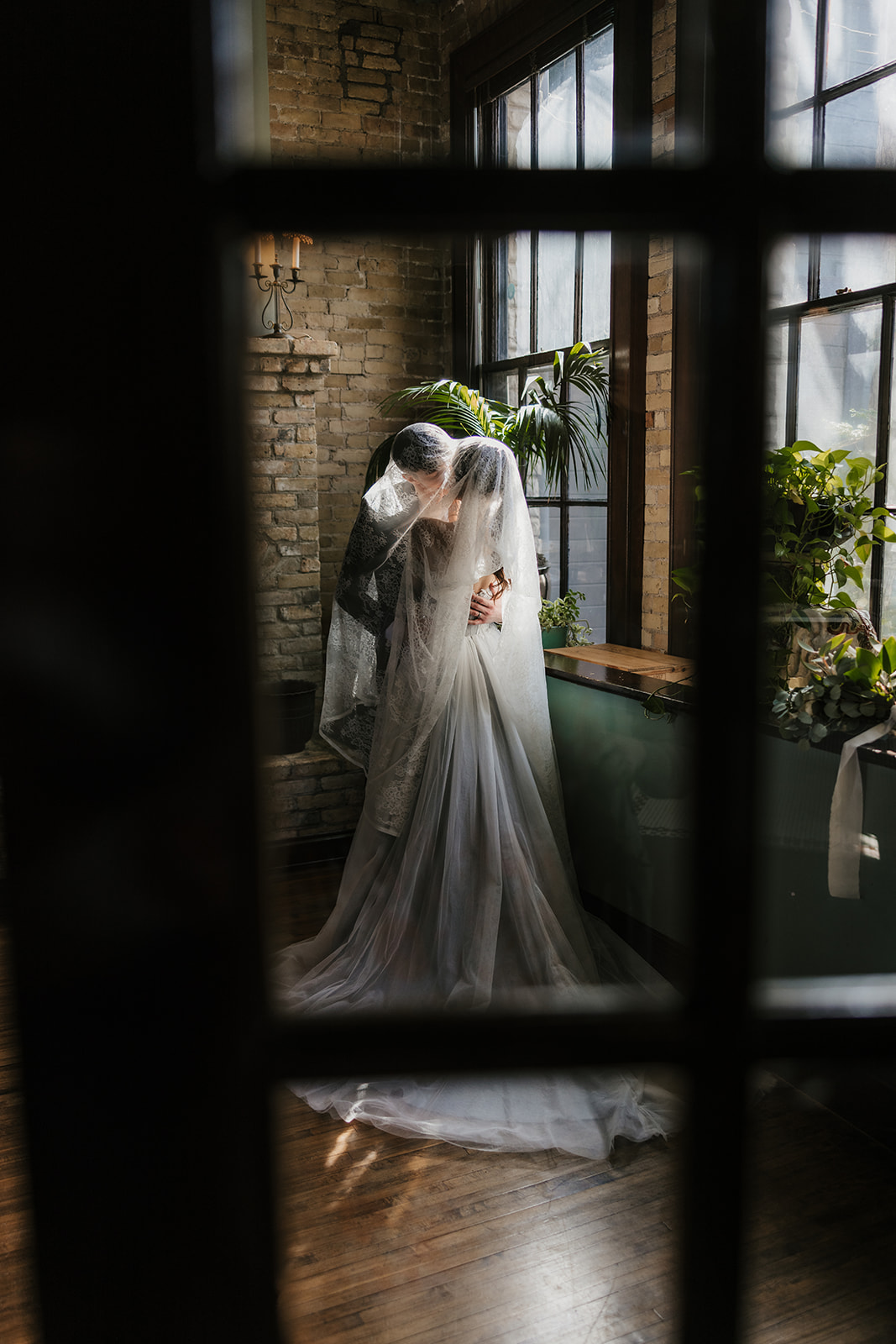 A bride and groom stand close together, gazing at each other in front of a window with sunlight streaming in, surrounded by greenery and brick walls for catholic wedding ideas