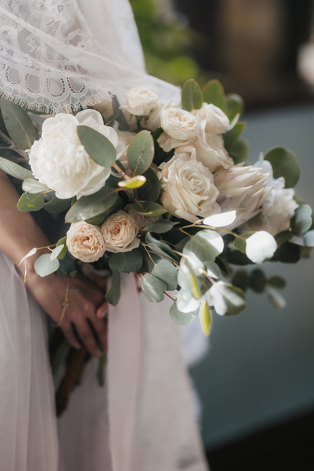 Person holding a bouquet of white and pale peach roses with green eucalyptus leaves, wearing a white lace garment.