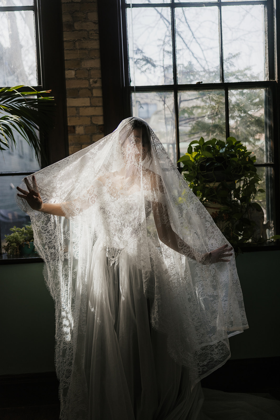 A person wearing a wedding dress and veil stands in front of large windows, illuminated by natural light.
